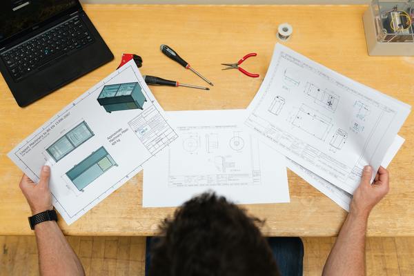 A imagem contém a vista aérea de uma mesa com papéis de projeto, algumas ferramentas e um notebook. Fazer uma pós em Engenharia abre várias possibilidades no mercado de trabalho.