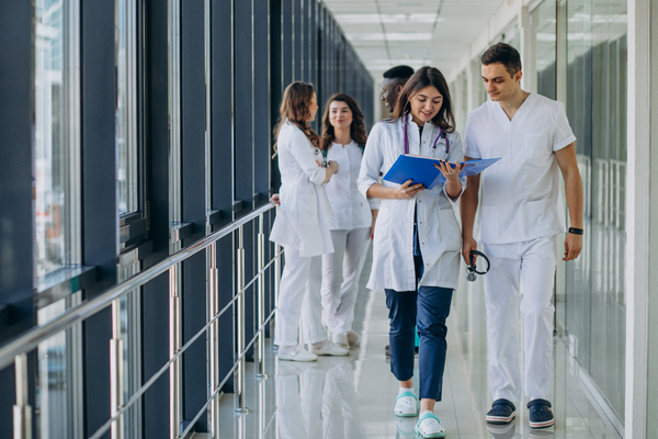 A imagem contém jovens médicos em um corredor. Três conversam ao fundo em desfoque, enquanto dois vão mais à frente tratando de assuntos relacionados à gestão de saúde.