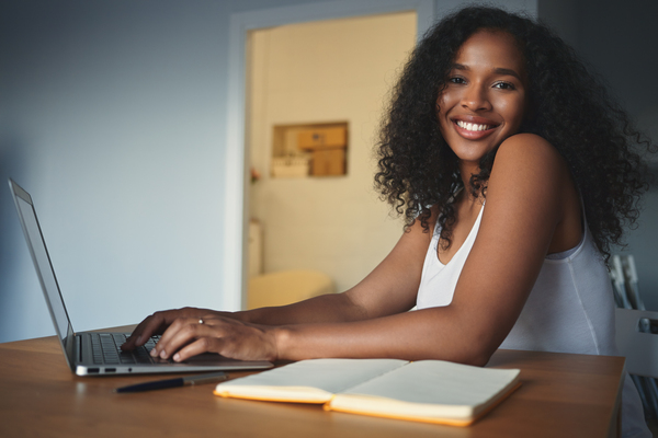 A imagem contém uma jovem estudante com seu notebook e livros abertos. A jovem, que sabe como organizar o tempo para estudar, está sorrindo.