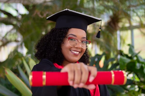 A imagem contém uma estudante com roupa de formanda, segurando o canudo e sorrindo.
