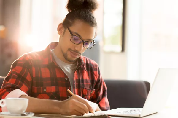 A imagem contém um jovem estudando para o vestibular de Medicina. Ele está sentado com o notebook aberto e faz anotações em um caderninho.